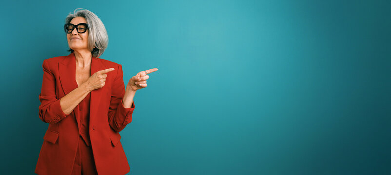 Senior businesswoman in a red suit and glasses pointing right, confidently presenting or indicating an area for text or product