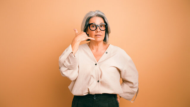 Mature woman wearing glasses and a natural shirt making a call me gesture, looking at the camera on an orange background