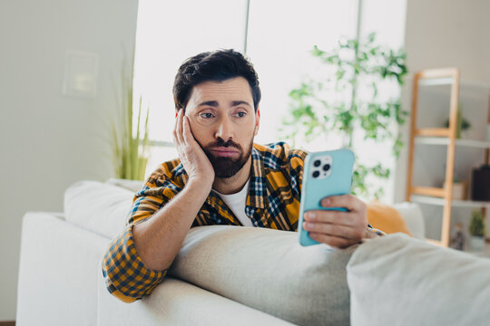 Man bored and scrolling phone on couch at home, casual young adult looking disappointed while using smartphone in bright living room