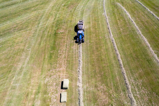 Aerial view of a blue tractor pulling a baler through a green field to harvest hay and create square bales in Chavannaz, Auvergne-Rhone-Alpes, France.