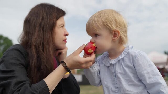 Adorable girl taking fruit from mom and biting it. Family and weekend concept. Happy childhood and healthy food. Cute baby girl child kid eating red apple seasonal fruit.