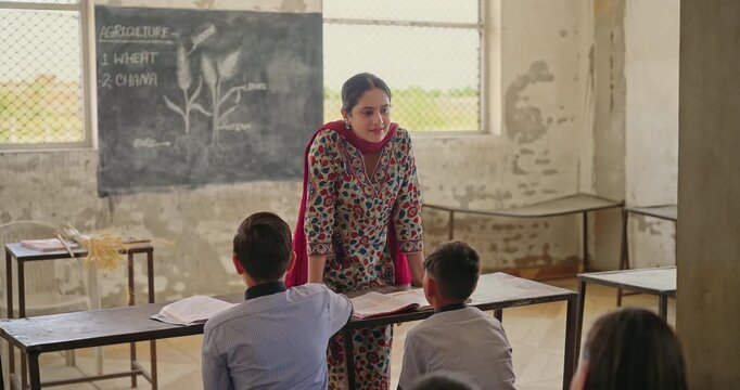 Indian young adult lady female teach lesson do study work indoor class room rural area place. Teen boy child sitting on bench wear uniform look tutor talk speak share story learn enjoy day desi life