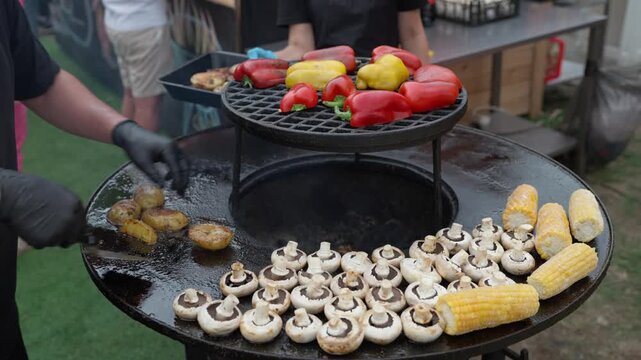 Round grill with fire inside. Champignon mushrooms, potatoes, and corn are cooked on the edge, while vegetables and bell peppers are cooked in the center. A cook removes cooked potatoes from the grill