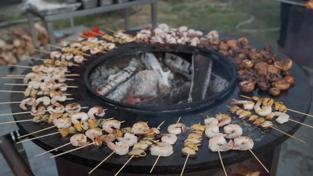 Appetizing seafood, shrimp, and mussels are grilled on skewers on a hot outdoor barbecue. Black flat grill, round shape, with a hole with a fire in center. 