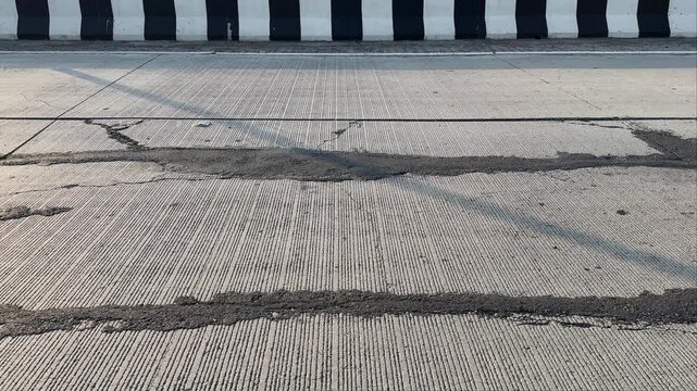 Tracking shot along a damaged concrete roadway featuring visible cracks, repaired patches, and textured pavement lines, with roadside safety barriers in the background under daylight conditions.
