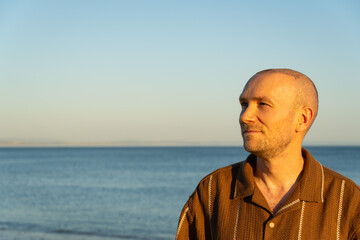 A profile portrait of a calm, pensive Caucasian man looking away into the distance, showing a healing scalp after a recent hair transplant procedure, standing by the ocean at sunset © maxterram