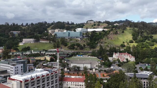 UC Berkeley campus with Beaux-Arts buildings, red-tile rooftops and construction crane beside lush green hillside and research labs under overcast skies, California, USA. Aerial drone view.
