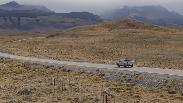 Crew cab pickup truck travels along Ruta Nacional 40 in Santa Cruz, Patagonia, Argentina, crossing open steppe beneath cloud-covered Southern Patagonian Andes, drone backward tracking parallax shot.