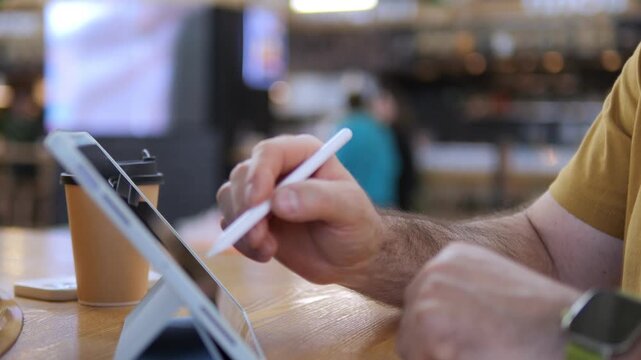 Person draws on a tablet with stylus in cafe. They wear yellow shirt and smartwatch. Coffee cup sits beside device on table. Scene shows calm, focused digital creativity moment