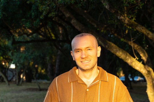 A portrait of a cheerful, broadly smiling Caucasian man displaying his healing scalp with scabs after a hair transplant operation, standing outdoors in a sunlit park.