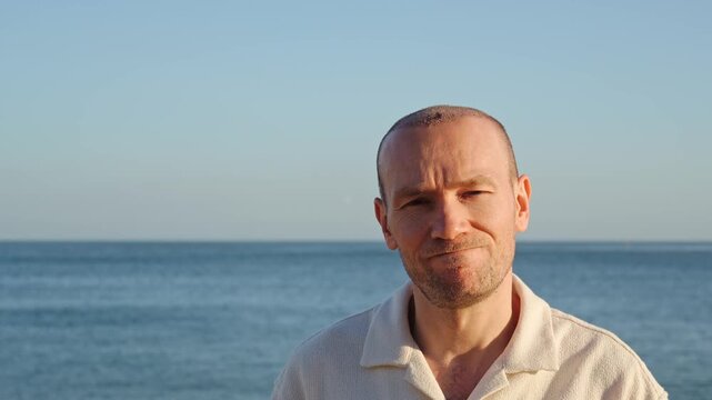 Mddle-aged man after a hair transplant procedure, looking at the camera with a hesitant expression head shaking on a sunny beach in Portugal during golden hour