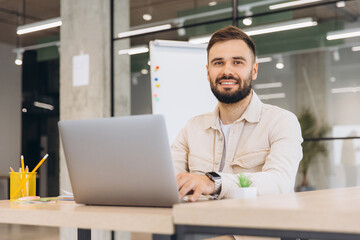Fototapeta premium Young professional man working on laptop in modern office, smiling at camera, showing confidence and success
