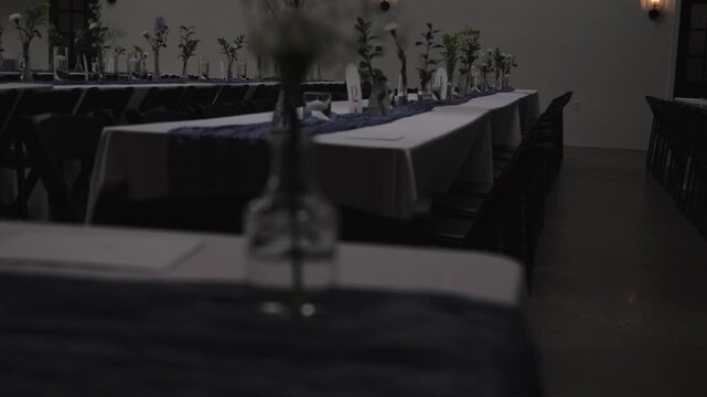 Long tables with minimal floral arrangements are set in a dimly lit reception hall. Suitable for event planning visuals or venue showcases.