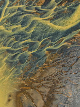 Aerial view of the intricate patterns of a braided glacial river with yellowish water flowing over dark volcanic sediment in Hella, Rangarthing ytra, Iceland.