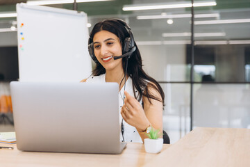 Obraz premium Indian Arabian woman smiling, wearing a headset, working on a laptop, engaging in customer service or virtual meeting