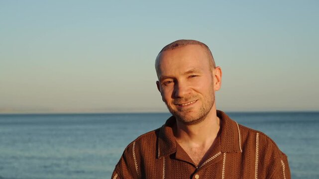 A portrait of a happy, smiling Caucasian man in a brown polo shirt, showing a healing scalp with visible scabs after a recent hair transplant procedure against a scenic sunset ocean background
