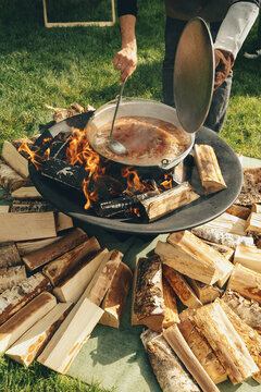 Cooking over a campfire in the countryside during the summer. A man stirs soup in a cauldron with a ladle.