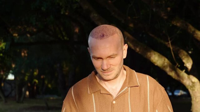 A portrait of a calm and content Caucasian man in a brown polo shirt, showing a healing scalp with scabs after a recent hair transplant procedure, standing in a sunlit park at sunset.