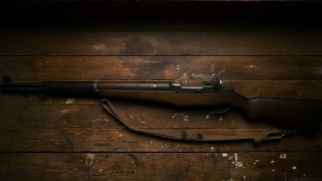 The iconic m1 garand rifle with a dark wooden stock and canvas sling rests on a weathered wooden surface in this high contrast shot showcasing the historical design of this classic military firearm.