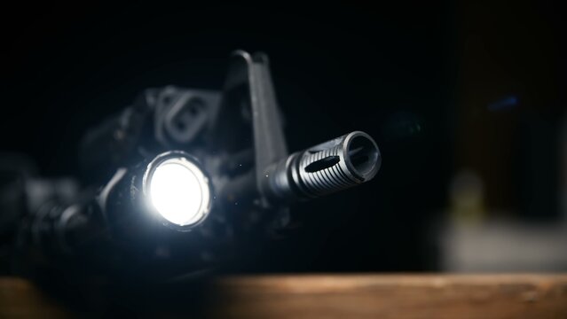 A black tactical rifle features a bright tactical flashlight mounted near the barrel and flash hider shown against a dark backdrop highlighting modern firearm attachments and specialized illumination 