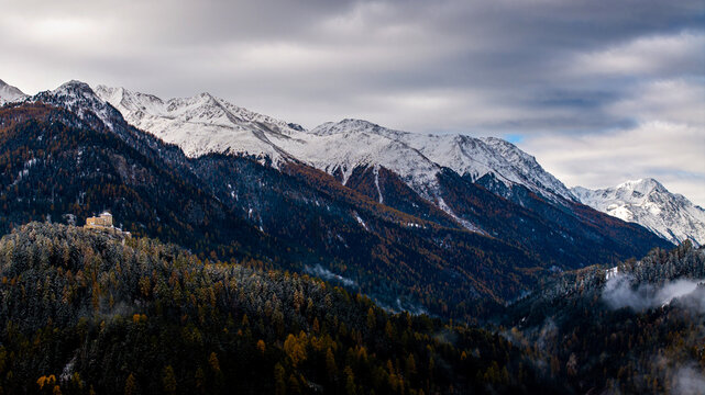 Aerial view of Schloss Tarasp perched on a hill surrounded by autumn forests and snow-capped mountain peaks under a cloudy sky in Scuol, Graubunden, Switzerland.