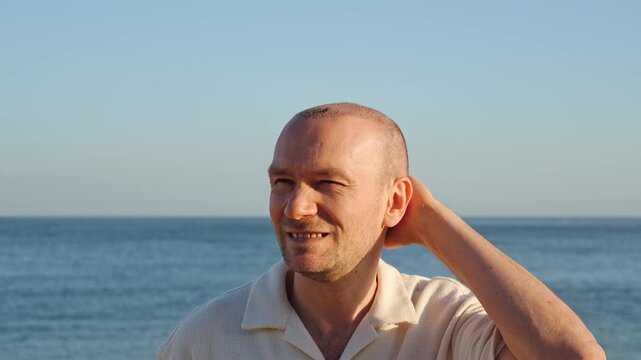 Portrait of a hesitant middle-aged man after a hair transplant procedure, scratching and rubbing his itchy shaved head while standing on a sunny beach in Portugal during beautiful golden hour