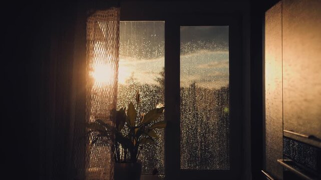 Golden sunset light shines through a rainy window with wet drops on a glass pane in a silent dark room. Sunset, rain, window, glass, drops, silent, light, mood, warmth, shadow, evening.