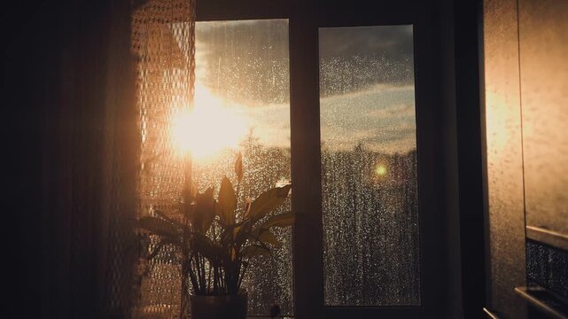 Golden sunset light shines through a rainy window with wet drops on a glass pane in a calm dark room. Sunset, rain, window, glass, drops, calm, light, mood, warmth, shadow, evening.