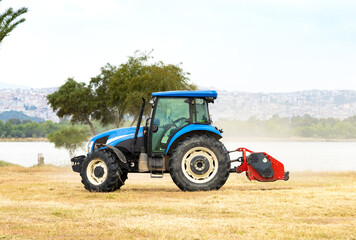 Obraz premium Modern blue agricultural tractor with mechanical equipment working on a dry grass field near a lake side with a distant city skyline under cloudy sky.