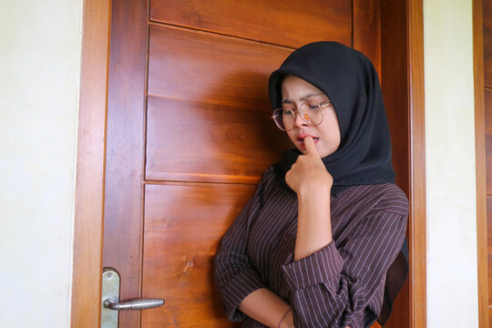 A worried Muslim woman wearing a black hijab and eyeglasses standing by a wooden door while biting her nail in anxiety or deep thought