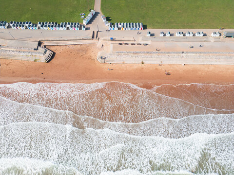 Aerial view of Preston Sands beach with white waves crashing on the red sand, colorful beach huts along the promenade and a green park area Paignton, Devon, United Kingdom.