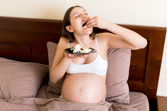 Hungry excited pregnant woman resting in bed is eating greedily a piece of cake and has a dirty mouth. Expecting mother can't stop eating