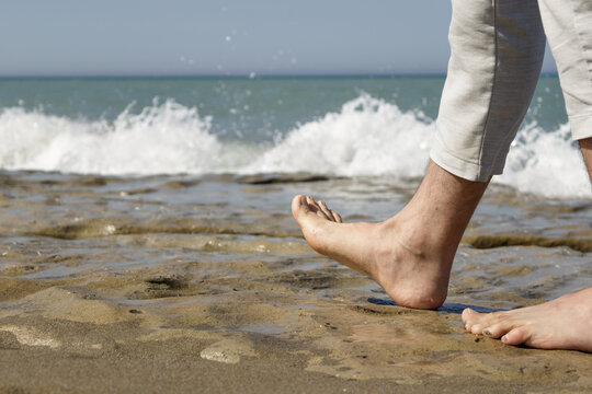 Bare feet walking on wet sandy beach with ocean waves at shoreline, person wearing rolled-up pants.