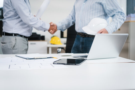 Two men shake hands in a bright office, one wearing a hard hat, suggesting a successful construction meeting, contract agreement, or project partnership.
