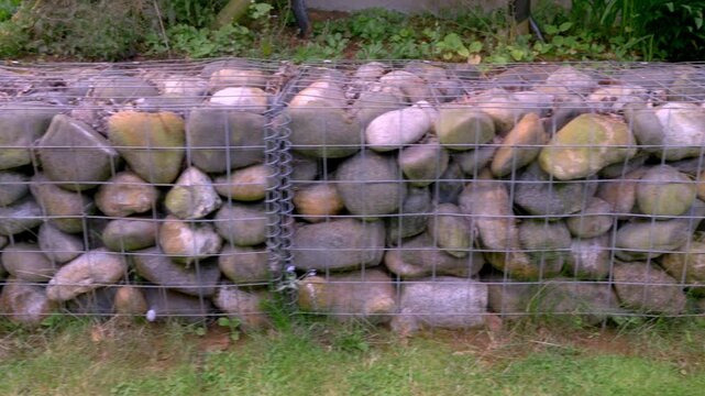 Closeup pan of large numbers of rounded stones or boulders inside horizontal wire cages, positioned next to a higher level of garden or yard, to act as a retaining wall to prevent soil from slipping.