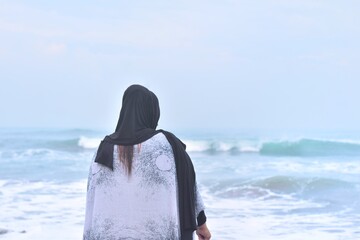 Asian woman wearing a black hijab and patterned white garment stands on the shore, observing the gentle waves of the ocean on a tranquil, overcast day. © Torao