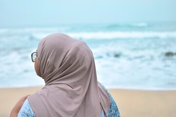 A thoughtful woman wearing a beige hijab and sunglasses stands on a sandy beach, looking towards the calm, blue-green ocean and gentle waves. © Torao
