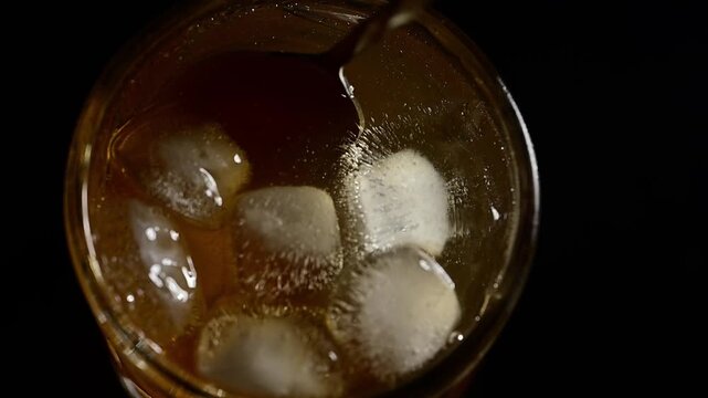 Top view of glass with alcoholic cocktail and ice cubes, bartender spoon stirring drink slowly on black background, mixing motion, cold refreshing beverage, professional bartending process