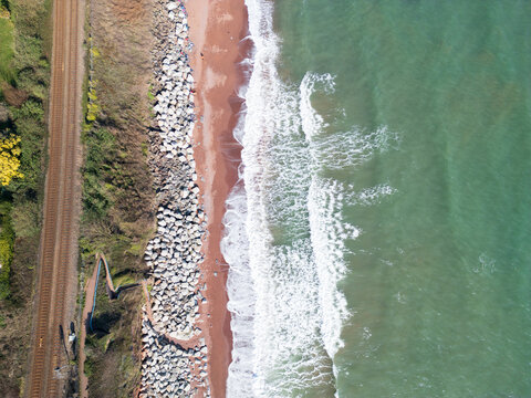 Aerial view of Hollicombe beach with railway tracks, rocky sea wall, and waves crashing on the shore in Torquay, Devon, United Kingdom.