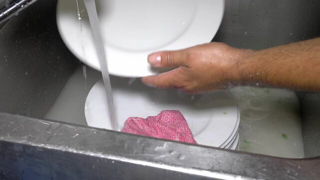 A kitchen worker rinses dirty plates under running water to remove leftover food residue, a careful preparatory step before loading them into the restaurant's industrial dishwasher.
