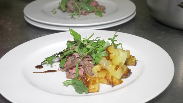 A chef carefully composes a plate with beef straccetti, fresh arugula and golden roasted potatoes, balancing colors and textures for an appealing presentation at the catered party event.