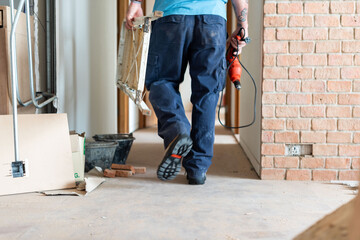 Person in work clothes carrying a drill and ladder in a construction area.
