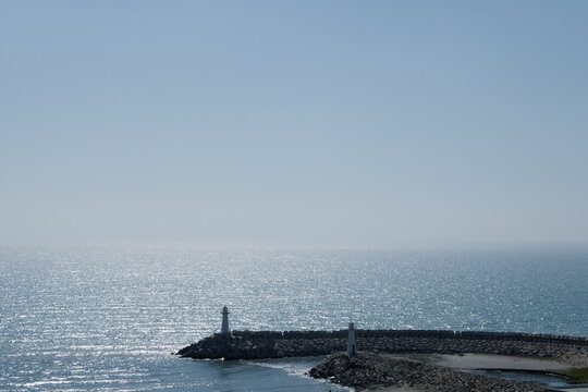Minimalist seascape with blue ocean and sky, lighthouse on breakwater or pier, calm horizon view, peaceful coastal scene on sunny day.