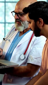 Indian male doctor using laptop while consulting young man patient as experienced physician discusses medical reports in modern Indian clinic OPD healthcare consultation