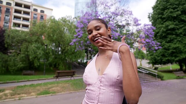 Playful tattooed young woman smiling and posing in a city park with jacaranda trees in bloom, waving and blowing a kiss while walking, radiating carefree happiness and joy
