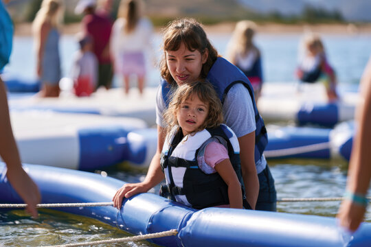 A mother embraces her young daughter while they stand on a floating mat, displaying a beautiful moment of bonding and adventure in a sunny water setting.