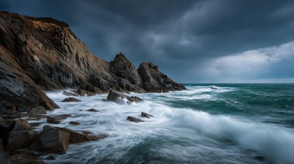 Fototapeta premium Rocky coastline with turbulent waves under a moody, cloudy sky, showcasing the power of ocean waters against jagged cliffs.