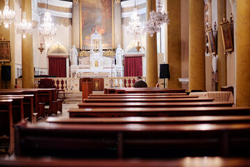 Naklejka premium Quiet church interior with wooden pews, chandeliers, altar, religious statues and a seated worshipper in warm light.