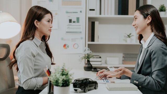Asian female car dealer explaining insurance policy or finance contract to client with red toy car on desk in modern showroom office.
