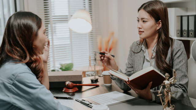 Professional female Asian lawyer holding law book and explaining legal contract details to a stressed client in corporate law firm office.
